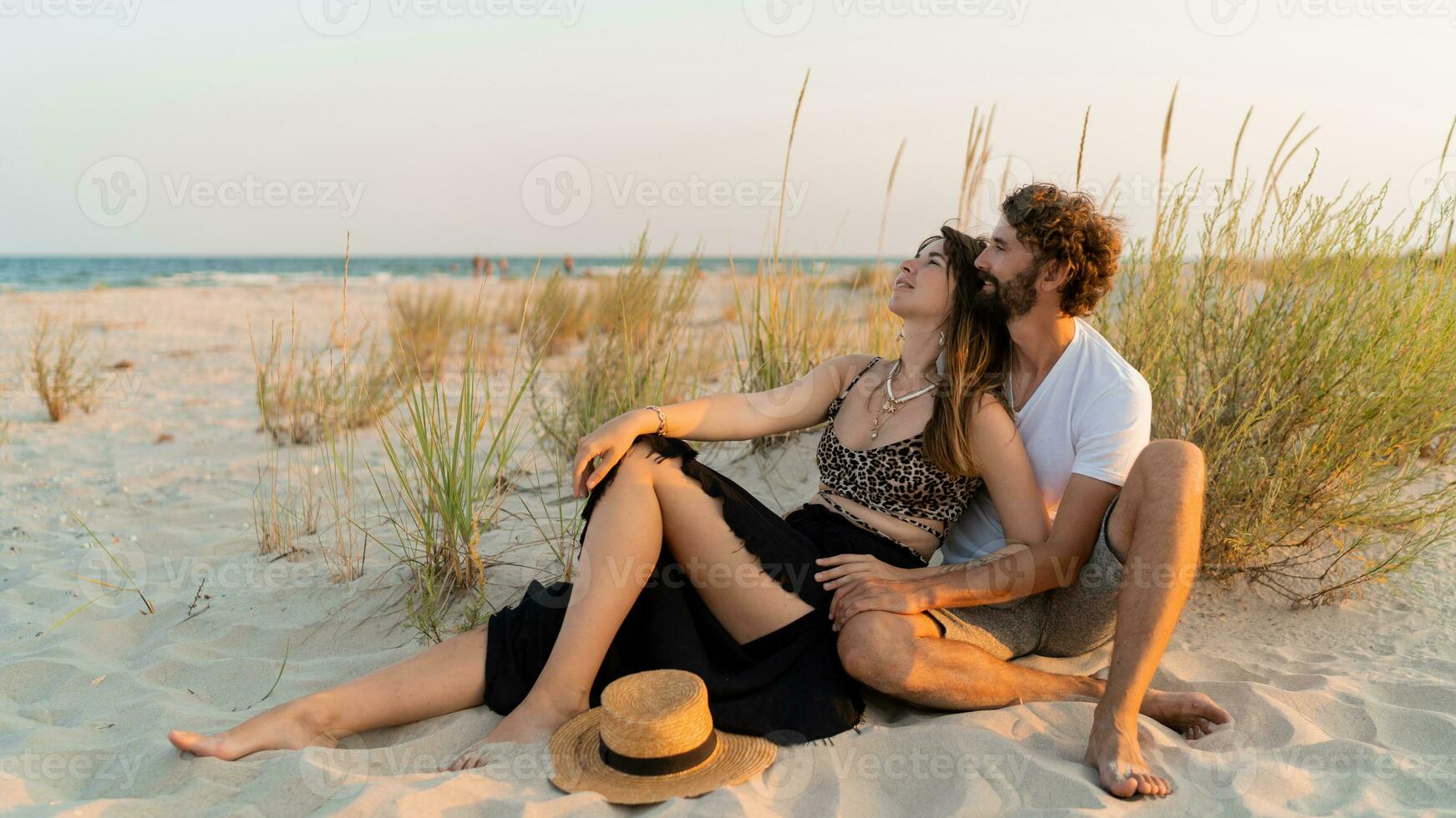 Stylish couple in love posing on the beach. Brunette woman in straw hat with her  boyfriend chilling in warm summer evening. photo
