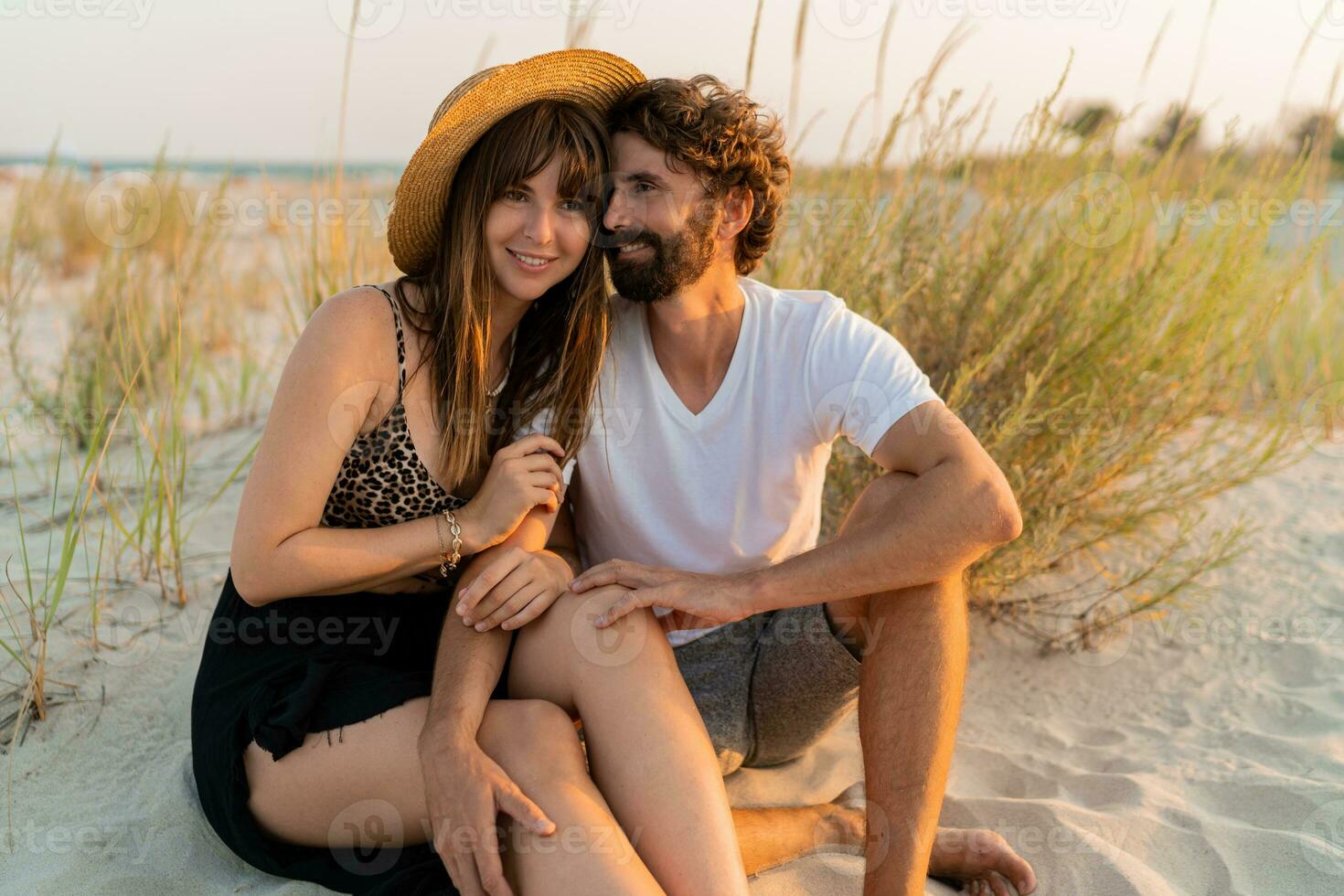 Traveling couple in love posing on the beach. Brunette woman in straw hat with her  boyfriend chilling in warm summer evening. photo