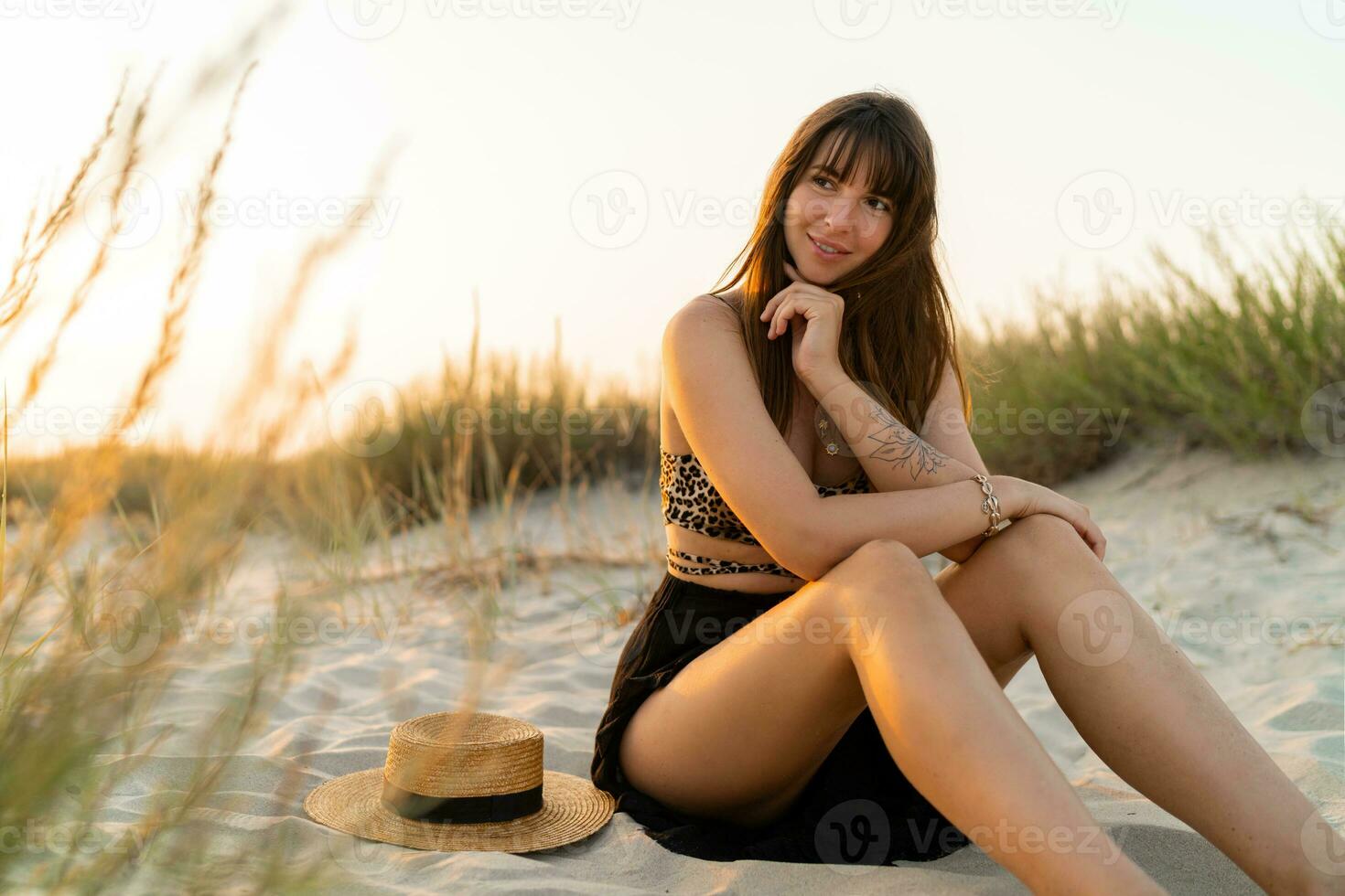 sexy mujer en elegante verano boho atuendo sentado en arena y disfrutando vacaciones en el playa. puesta de sol colores. foto