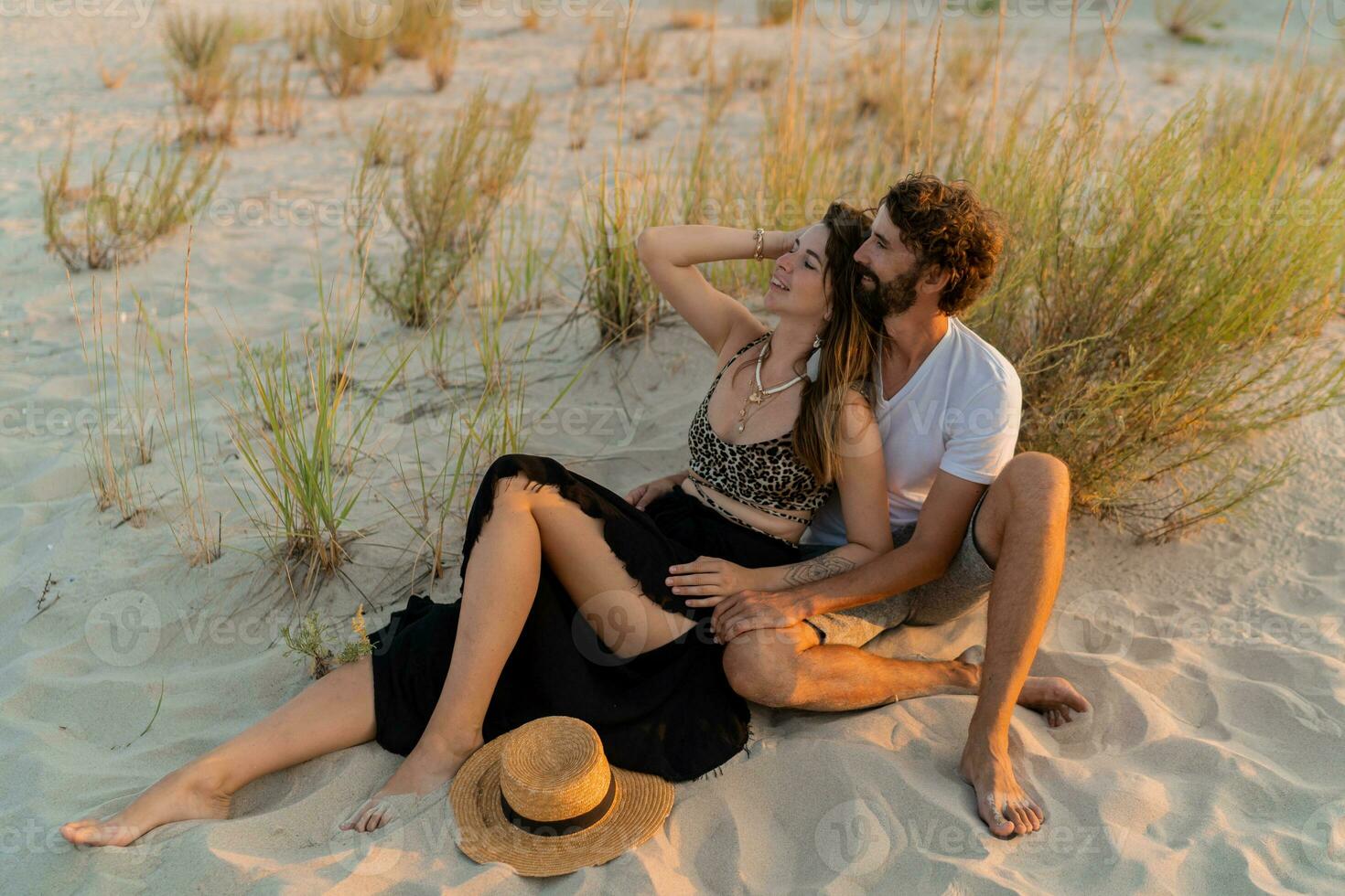 Traveling couple in love posing on the beach. Brunette woman in straw hat with her  boyfriend chilling in warm summer evening. photo