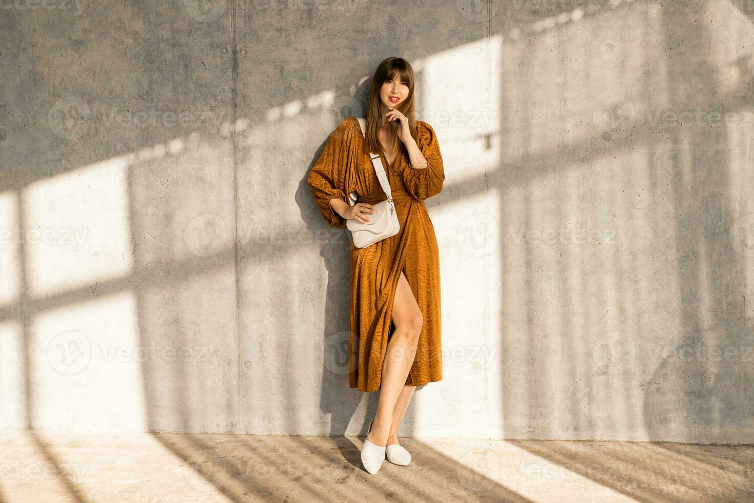 Indoor studio photo of elegant brunette woman in stylish dress standing over urban concrete wall.