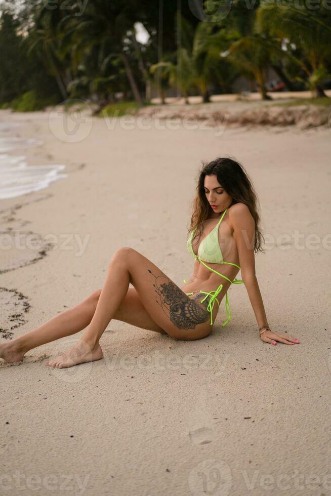 Beautiful tanned woman sitting  and sunbathing at the beach. Fashionable  slim  girl.  Outdoor photo. Summer vibes. Stylish swimwear. photo