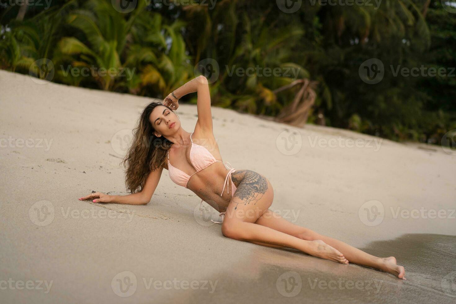 Beautiful tanned woman sitting  and sunbathing at the beach. Fashionable  slim  girl.  Outdoor photo. Summer vibes. Stylish swimwear. photo