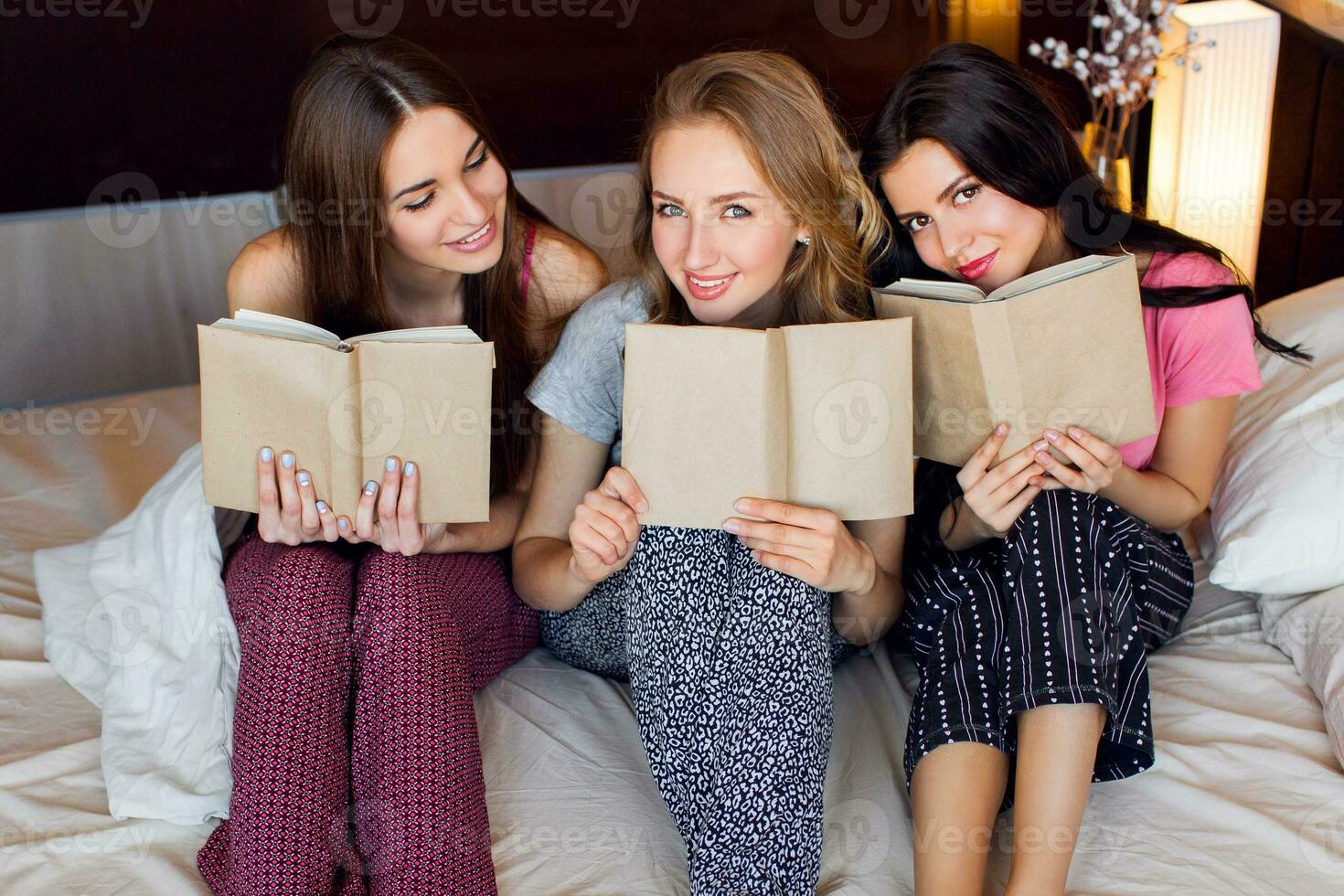 Lifestyle  image of three best friends in pajamas  reading  books, studying  and conversations  in bedroom.   Group of students  doing homework together at home. Cozy warm colors. Emotional faces. photo