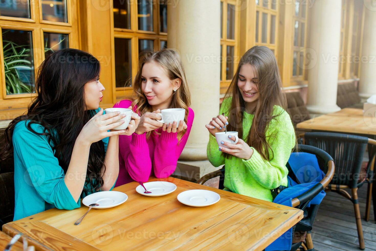 Close up lifestyle portrait of three beautiful  young  women sitting in cafe and enjoying  hot tee . Wearing bright neon yellow , pink and blue stylish  sweater .Holidays, food and tourism concept . photo