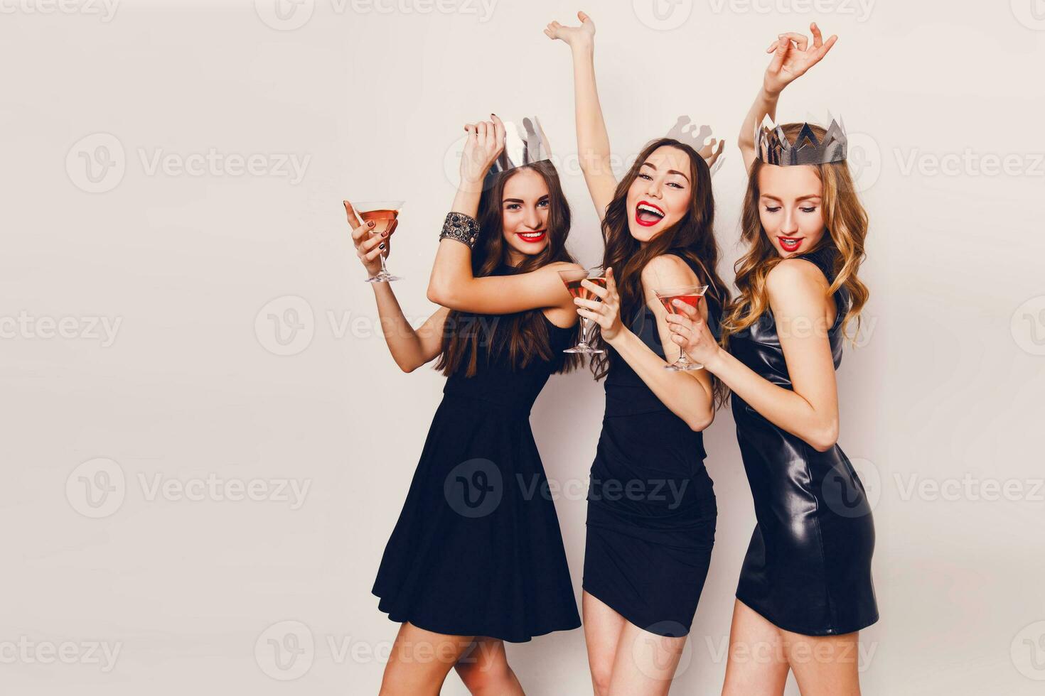 Three beautiful elegant women celebrate hen-party and drinking cocktails. Best friends wearing black  stylish evening dress,  high heel shoes ,crown on head .Bright make up, red lips. Inside. photo