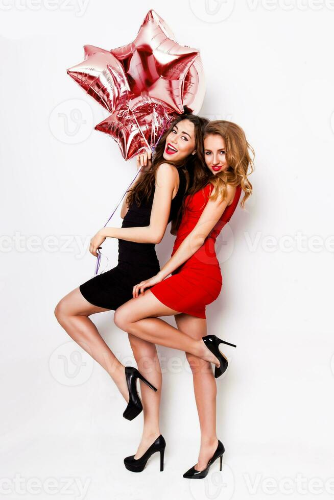 Two beautiful elegant women with red lips in evening black and red dress having fun. One keeping red stars balloons in her hand and smiling. Two woman at the party. Inside. White background. photo