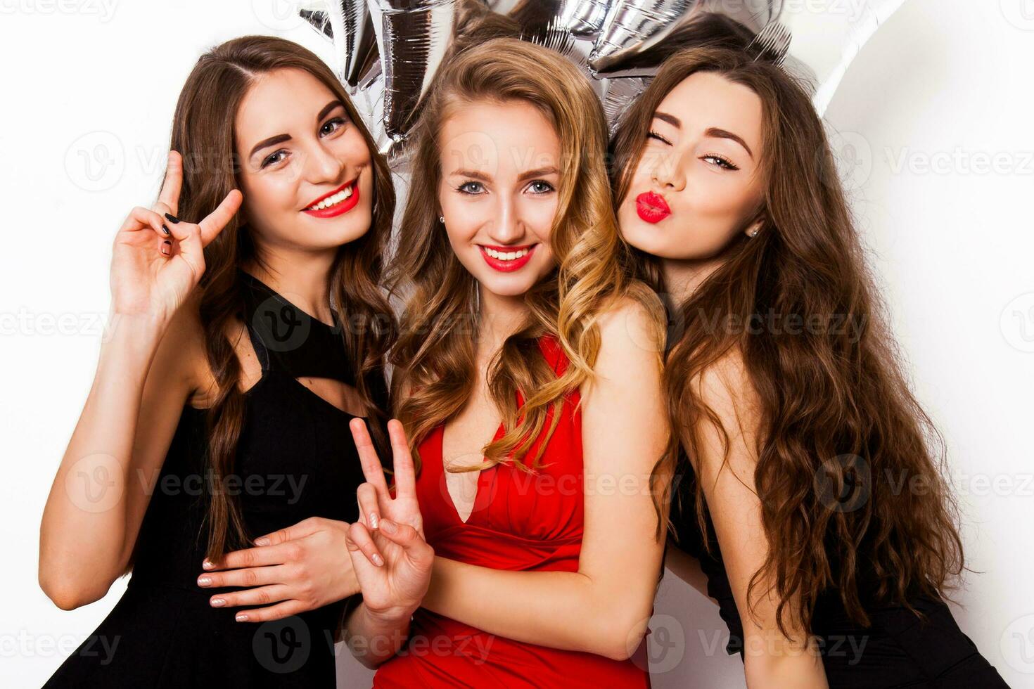 Three best friends celebrate  birthday indoor wearing elegant evening dress and have bright make up. Girls hugging and Showing signs with their hands. Looking at camera and smiling. Inside. photo