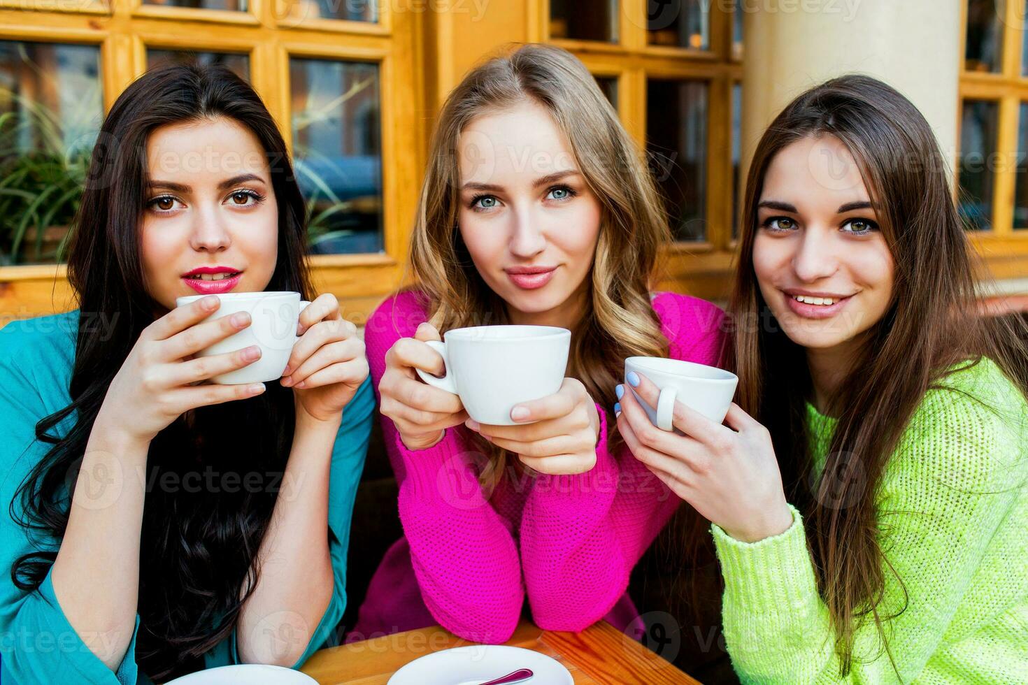 Close up lifestyle portrait of three beautiful  young  women sitting in cafe and enjoying  hot tee . Wearing bright neon yellow , pink and blue stylish  sweater .Holidays, food and tourism concept . photo