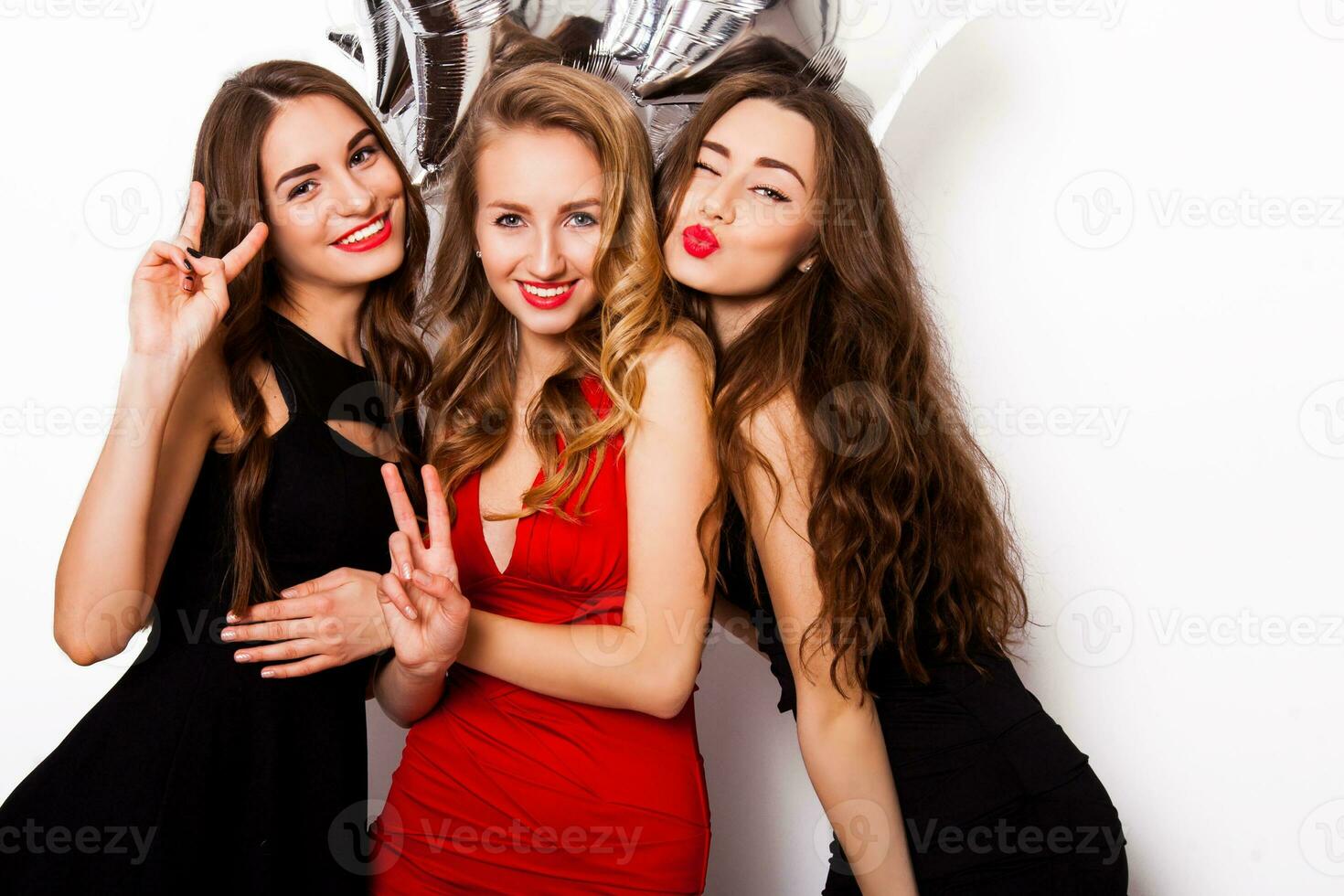 Three best friends celebrate  birthday indoor wearing elegant evening dress and have bright make up. Girls hugging and Showing signs with their hands. Looking at camera and smiling. Inside. photo