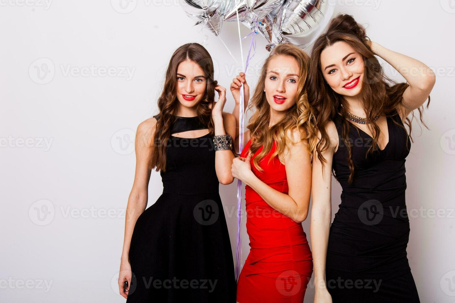 Three best friends celebrate  birthday indoor wearing elegant evening dress and have bright make up. Girls hugging and Showing signs with their hands. Looking at camera and smiling. Inside. photo