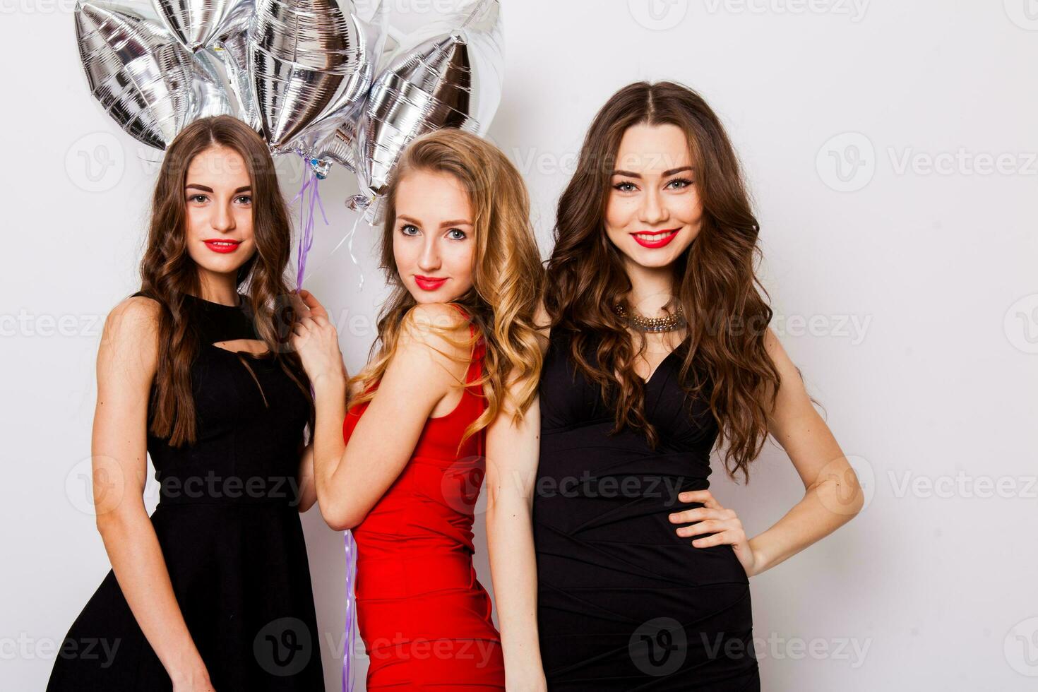Three best friends celebrate  birthday indoor wearing elegant evening dress and have bright make up. Girls hugging and Showing signs with their hands. Looking at camera and smiling. Inside. photo