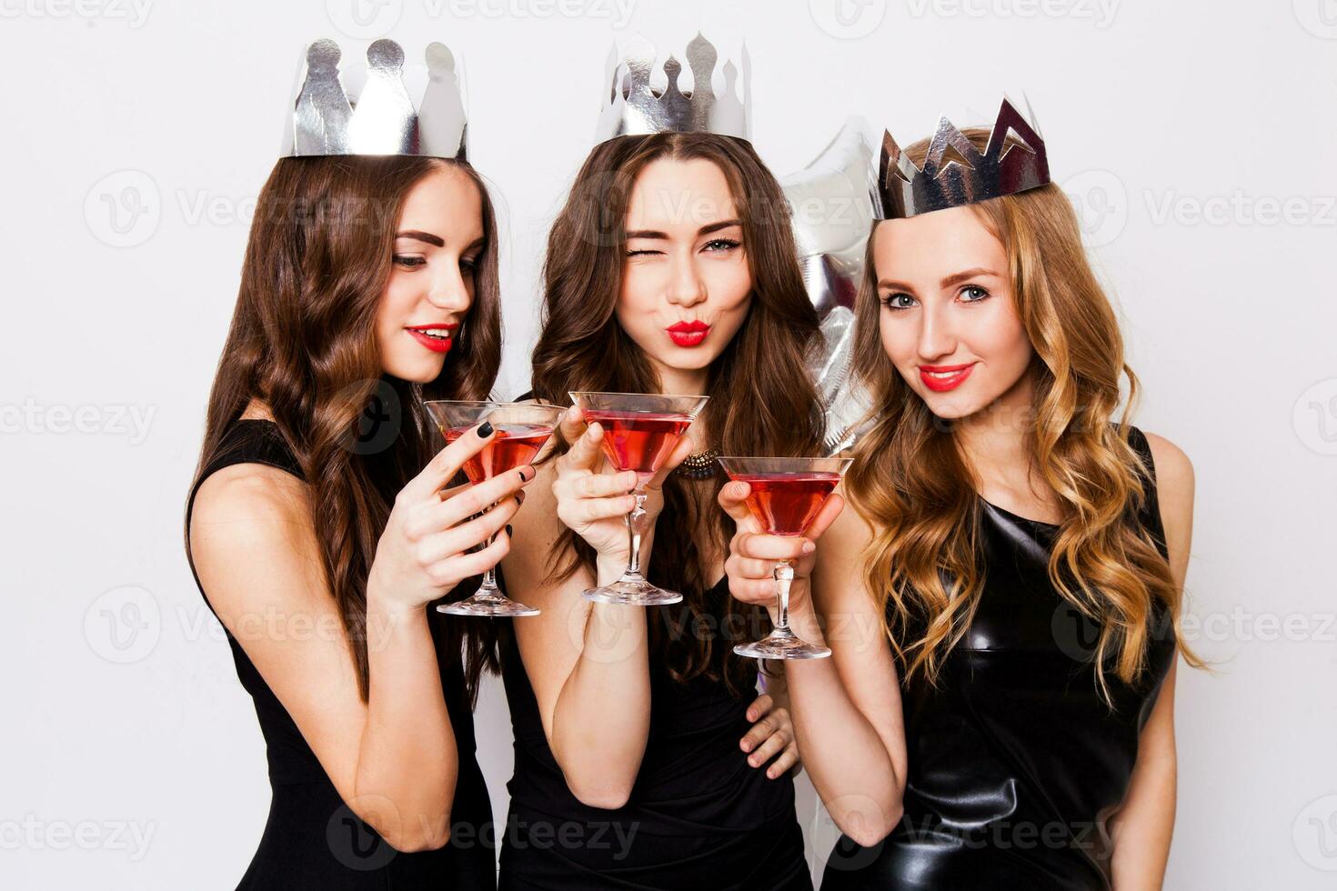Three beautiful elegant women celebrate hen-party and drinking cocktails. Best friends wearing black stylish evening dress, high heel shoes ,crown on head .Bright make up, red lips. Inside. photo