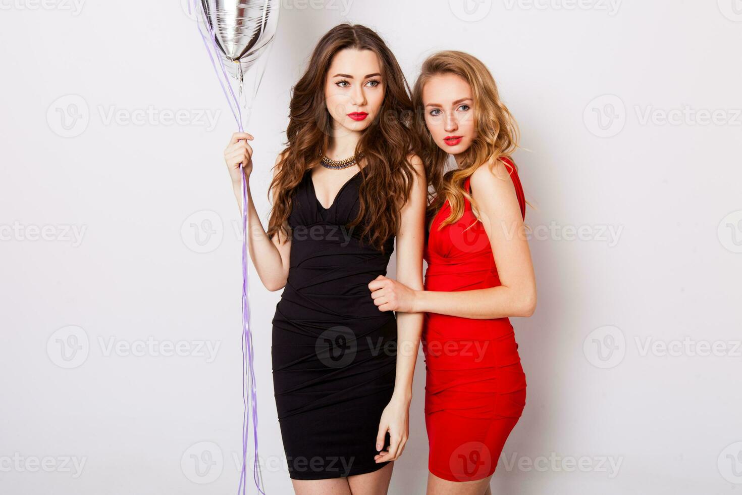 Two beautiful elegant women with red lips in evening black and red dress having fun. One keeping red stars balloons in her hand and smiling. Two woman at the party. Inside. White background. photo
