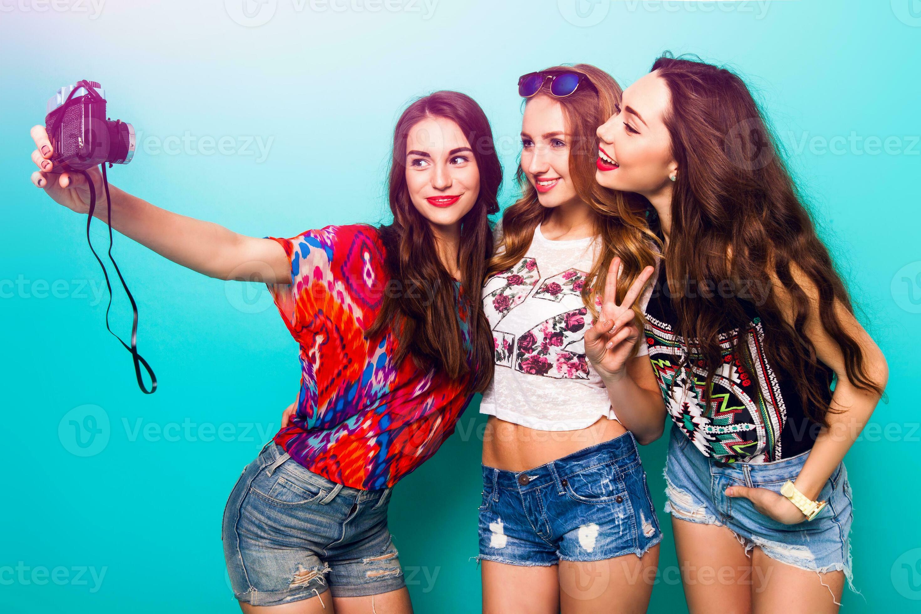 Three pretty sexy girls taking self portrait . Wearing bright summer  stylish outfit and jeans shorts . They smiling and do hand sings against  blue wall. 32631781 Stock Photo at Vecteezy