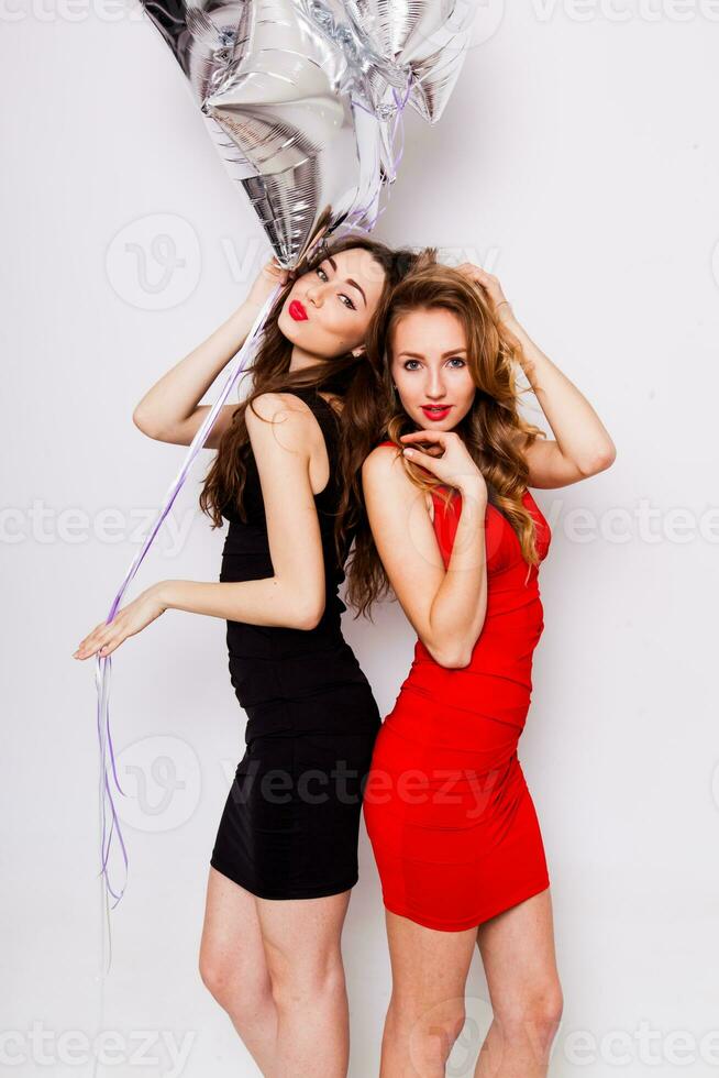 Two beautiful elegant women with red lips in evening black and red dress having fun. One keeping red stars balloons in her hand and smiling. Two woman at the party. Inside. White background. photo