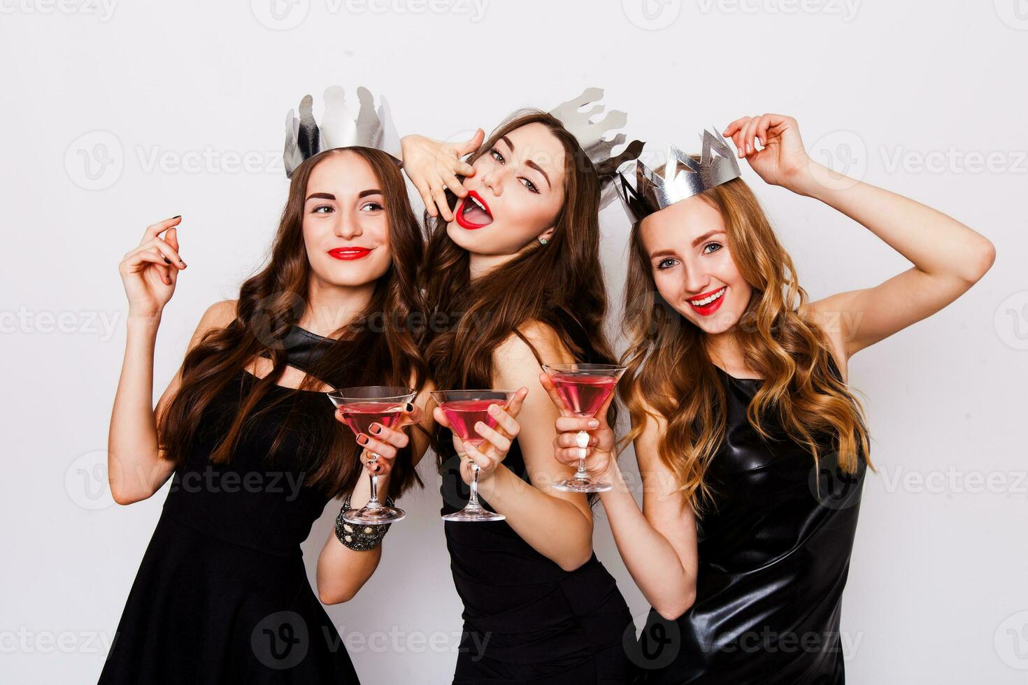 Three beautiful elegant women celebrate hen-party and drinking cocktails. Best friends wearing black  stylish evening dress,  high heel shoes ,crown on head .Bright make up, red lips. Inside. photo