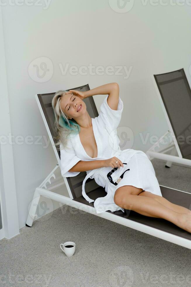Beautiful blond woman in white robe chilling  on deck chair in luxury hotel. Enjoing morning and drinking coffee. photo
