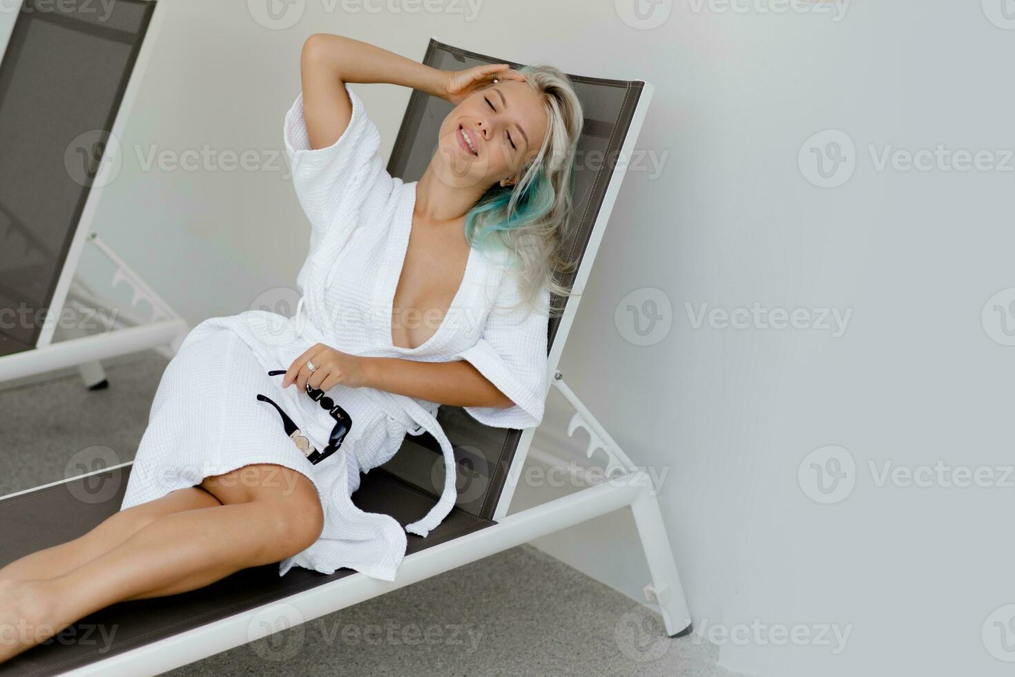 Beautiful blond woman in white robe chilling  on deck chair in luxury hotel. Enjoing morning and drinking coffee. photo