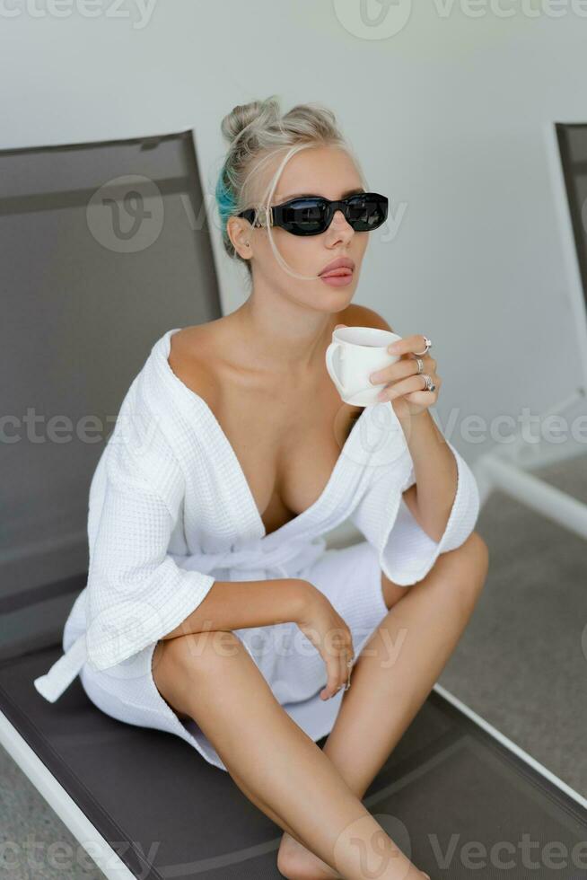 Summer photo of pleasure blond woman  in stylish sunglasses   and  white robe drinking cup of coffe in early morning.
