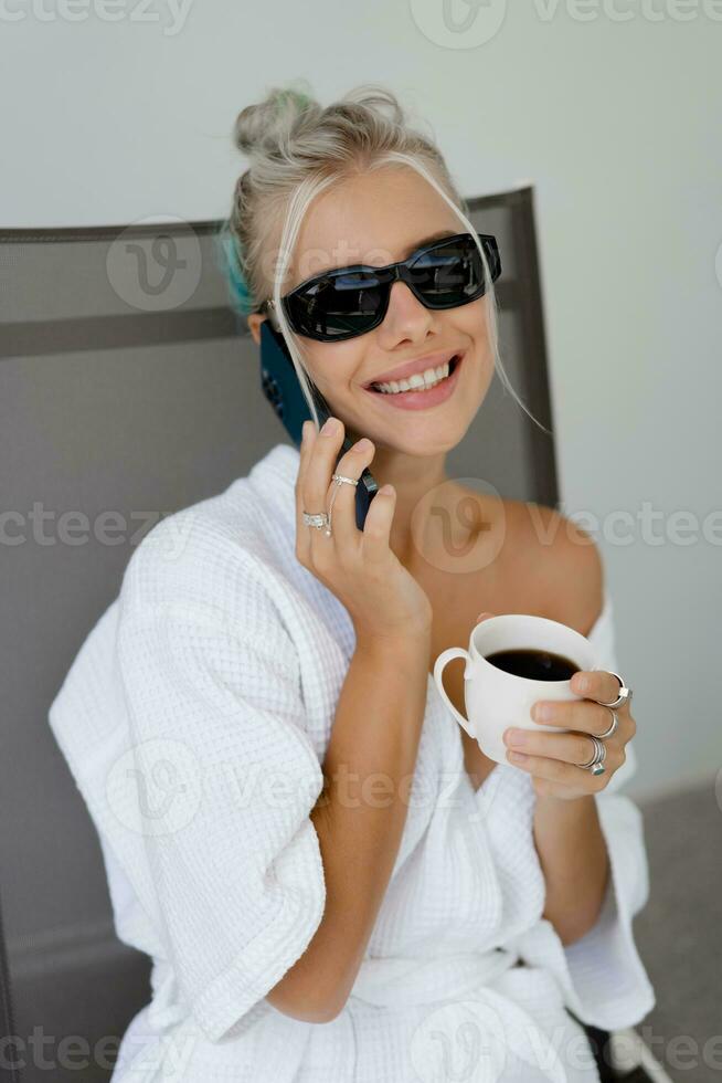 Close up portrait  of smiling  blond woman  in stylish sunglasses   and  white robe drinking cup of coffe in early morning. Resting in luxury hotel. photo