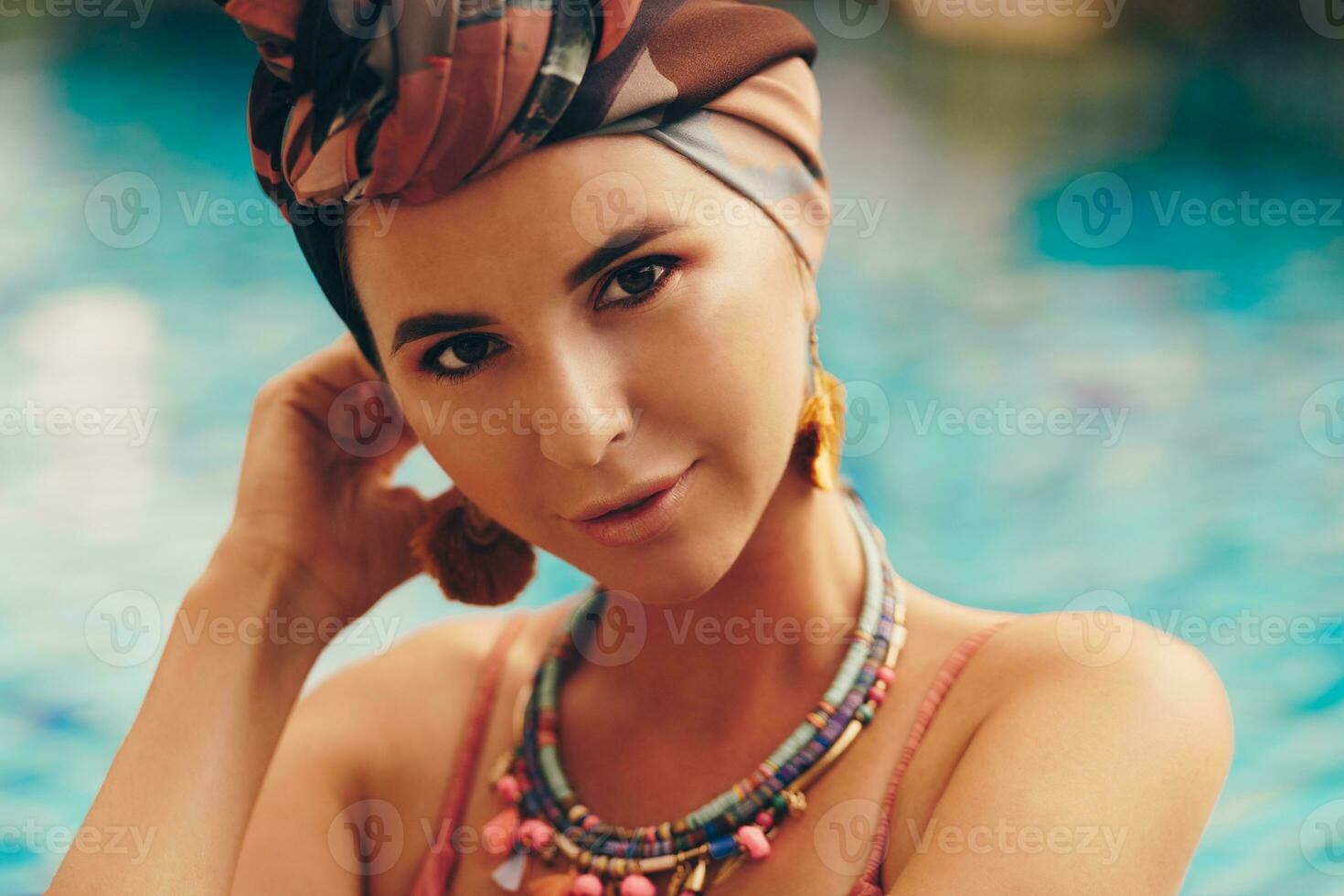 Woman with bright make up in bohemian outfit, turban, ethnic earrings, necklace and bracelets posing in swimming pool. photo