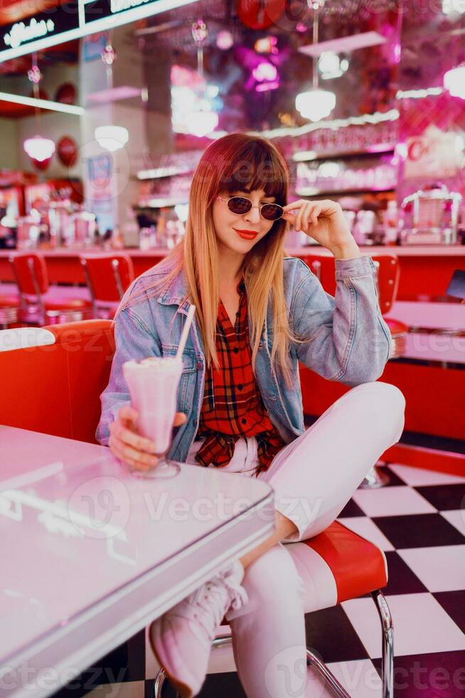 Fashionable  girl in  jeans jacket sitting   in t retro American cafe. Lovely smiling woman enjoying sweet milk shake . Wearing white sneakers. photo