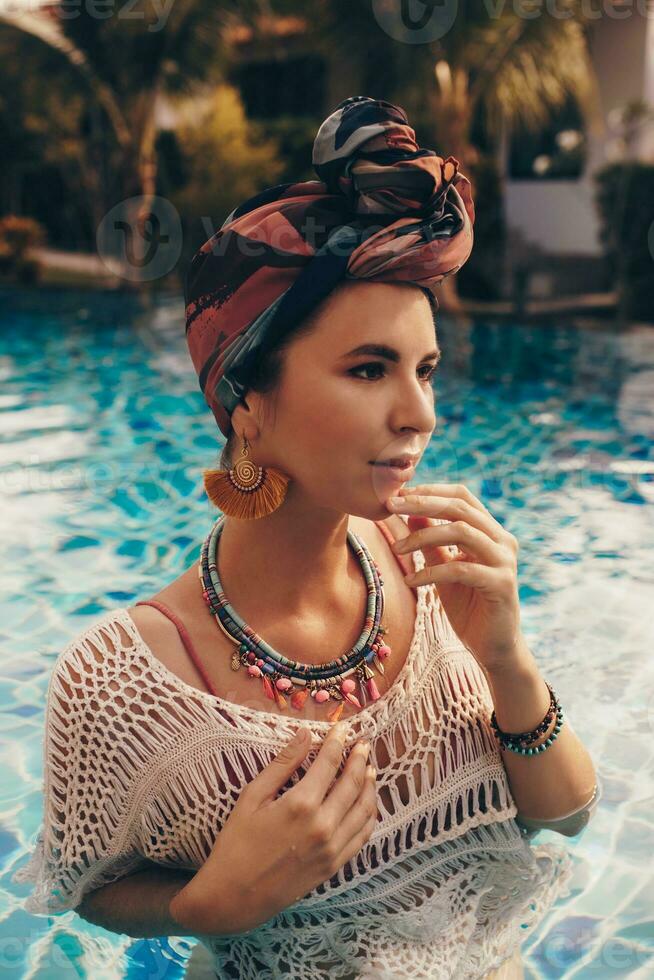 Woman with bright make up in bohemian outfit, turban, ethnic earrings, necklace and bracelets posing in swimming pool. photo