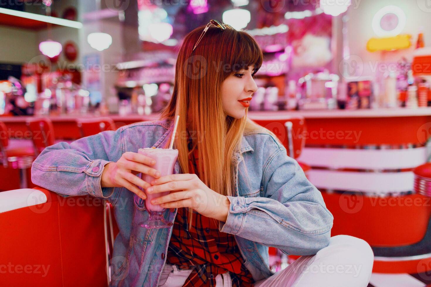 Fashionable  girl in  jeans jacket sitting   in t retro American cafe. Lovely smiling woman enjoying sweet milk shake . Wearing white sneakers. photo