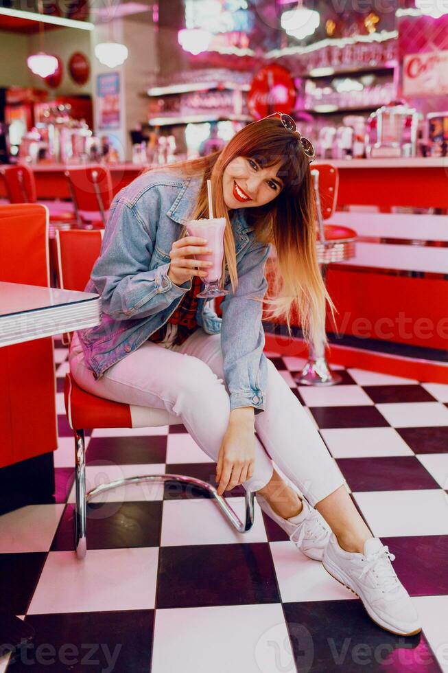 Full length  portrait of  long haired brunette woman with bright make up and red lips enjoying  day off  in stylish vintage American cafe  with neon lifts and red furniture. Drinking milk shake. photo