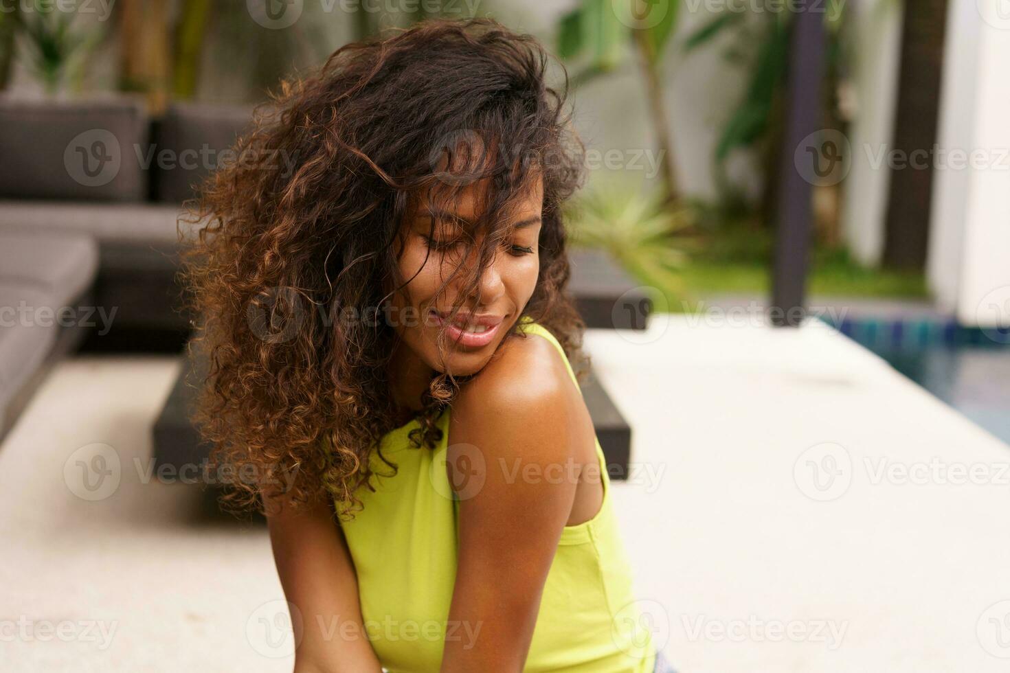 A perfect summer moment unfolds as a beautiful young African American woman, her curly hair framing her face, relaxes on a terrace of a tropical villa.. photo