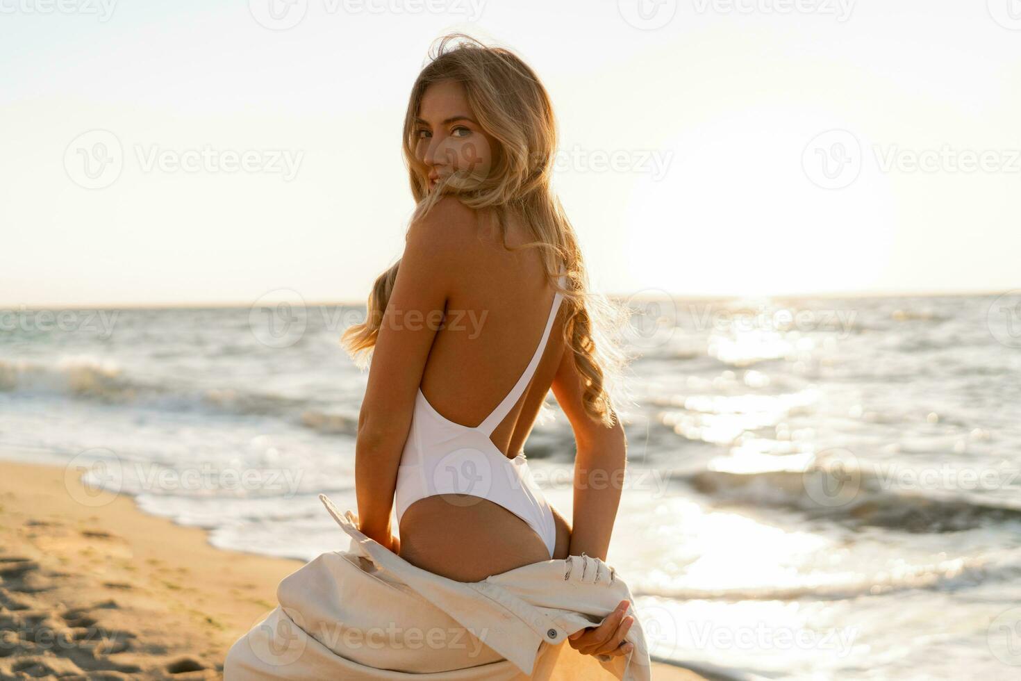 seductor rubio mujer en sexy trajes de baño posando en el playa en puesta de sol ligero. perfecto ondulado pelos, bronceado desnatar cuerpo. verano tropical humor. foto
