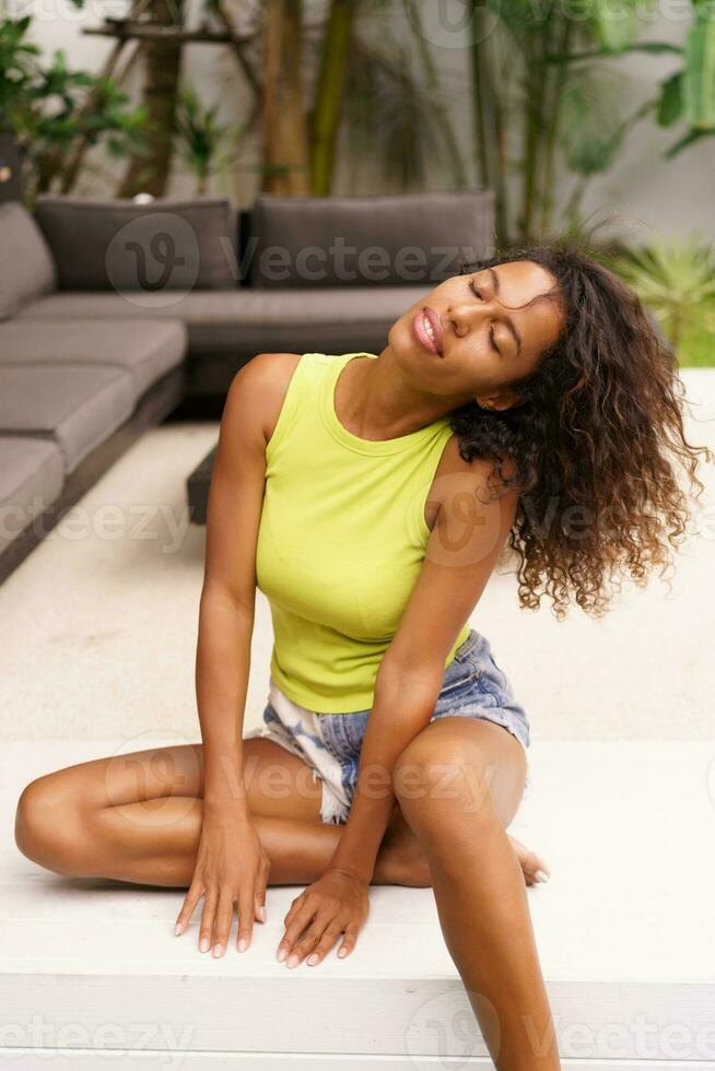 A perfect summer moment unfolds as a beautiful young African American woman, her curly hair framing her face, relaxes on a terrace of a tropical villa. . photo