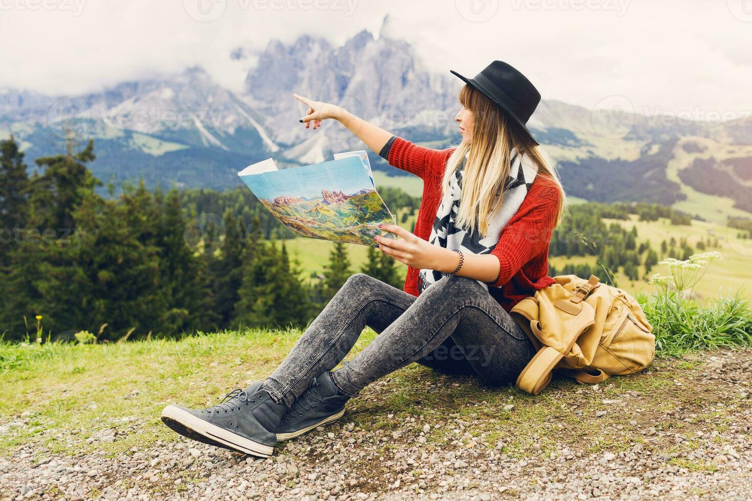 Traveler young woman  with backpack and hat sitting on grass  and searching right direction on map near amazing mountains and forest view. Shows a finger to the top. Soft toned colors. photo