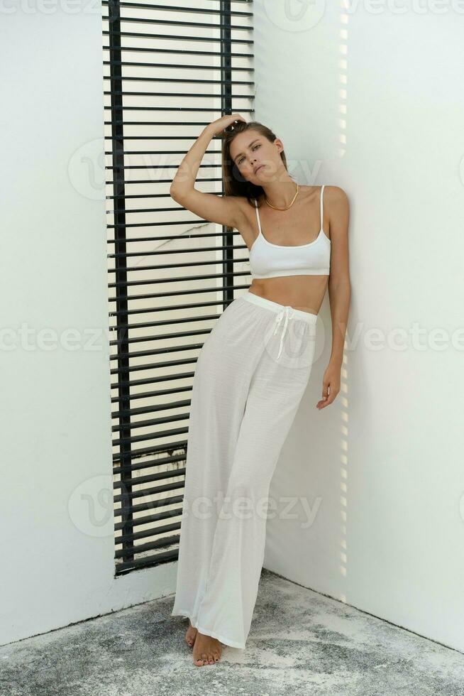 Fashion  photo of stylish brunette woman in white  homewear. Linen pants and crop top. Posing on terrace. Stylish modern interior. Full lenght.