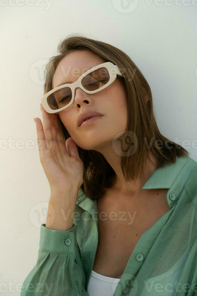 Close up portrait of stylish  woman in  sunglasses posing over wite background.  Casual outfit. Green linen blouse . white crop top and pants. photo