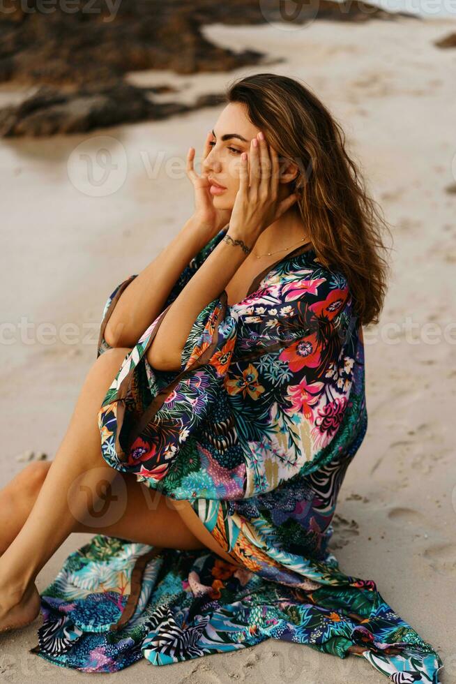 Geourgeous sensual  woman in pink swimsuit, and boho kimono  with tropical print  posing on  the beach at sunset. photo