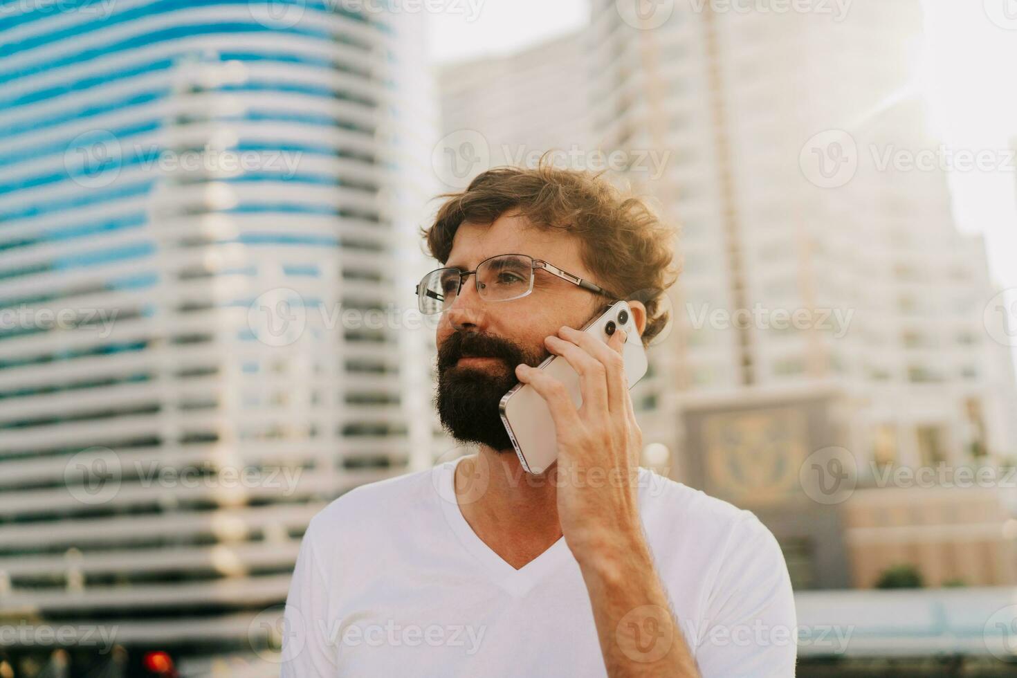 Handsome man with beard  talking by mobiyle phone while walking in big modern city with skyscrapers. Outdoors at sunset . Wearing white t shirt. photo