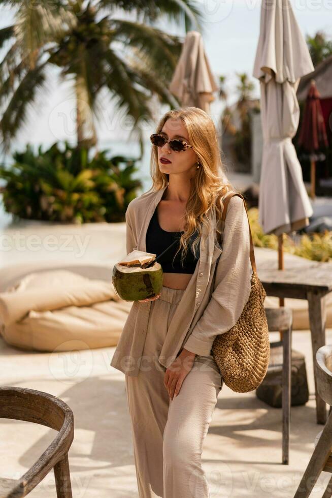 Pretty blond woman in stylish sunglasses dinking fresh coconut and posing in stylish beach club. Wearing summer linen costume. photo