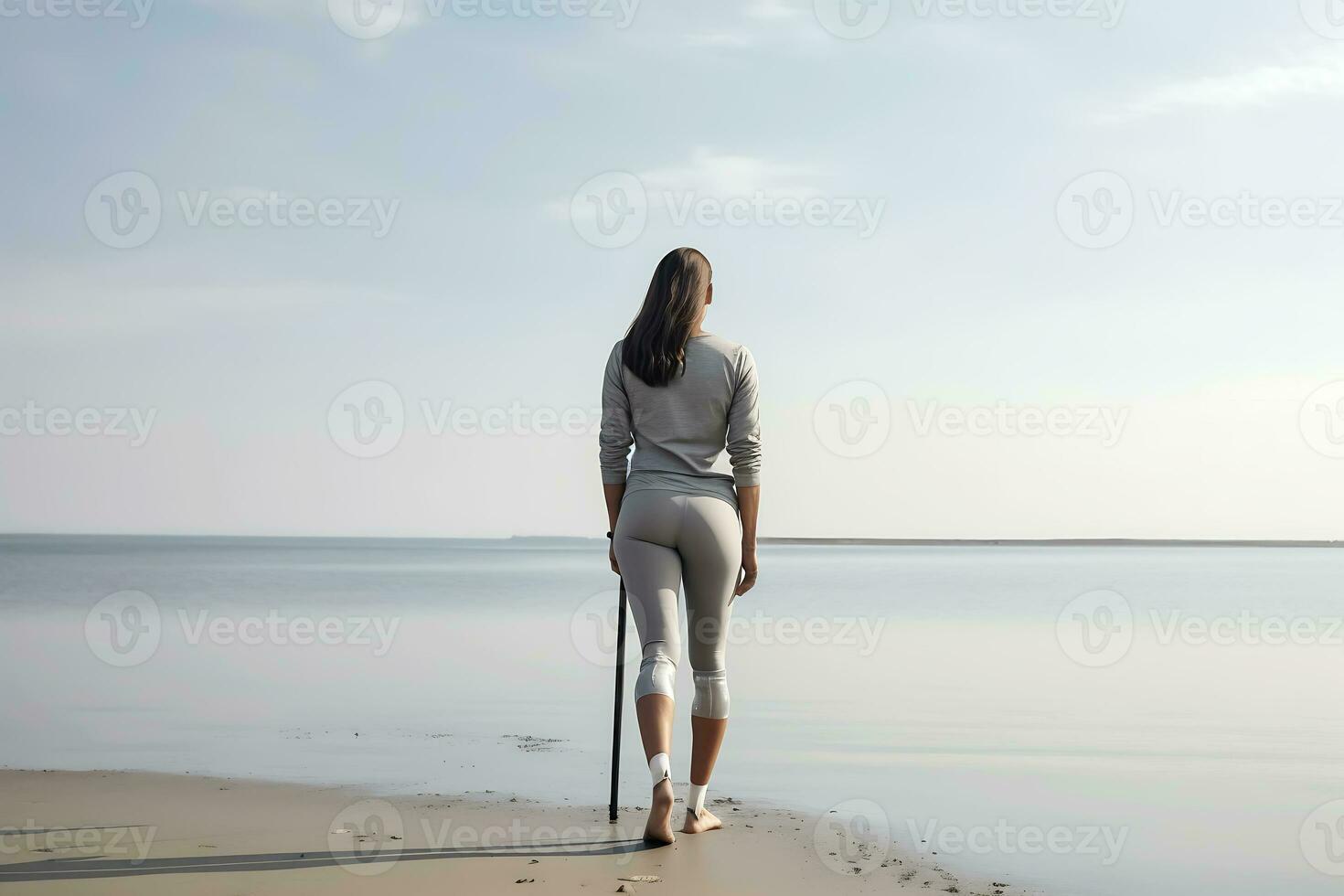 A young disabled woman with a cane with prosthetic legs in a gray tracksuit poses against the backdrop of the sea in sunny weather. Woman posing with her back to the camera photo