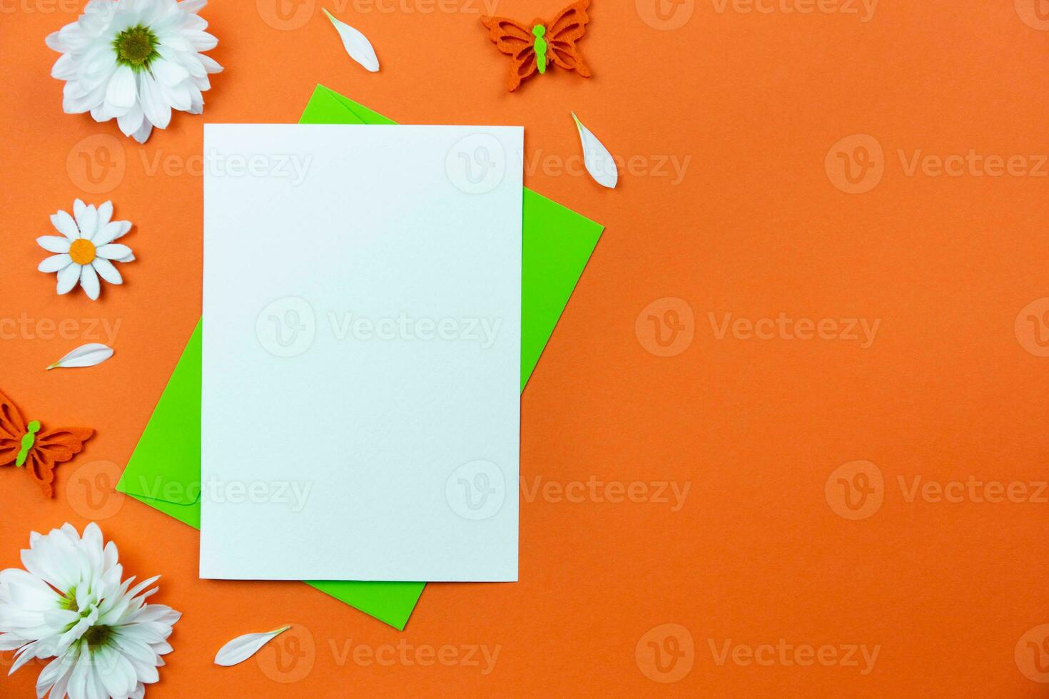 Blank greeting card mockup on colorful orange background with white daisy flowers and butterflies decoration. Spring flat lay composition. Top view. Copy space. photo