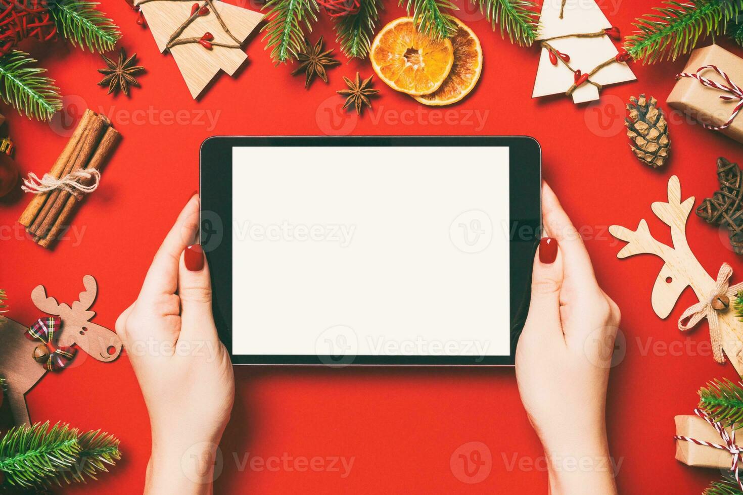 Top view of woman holding tablet in her hands on red background made of Christmas decorations. New Year holiday concept. Mockup photo