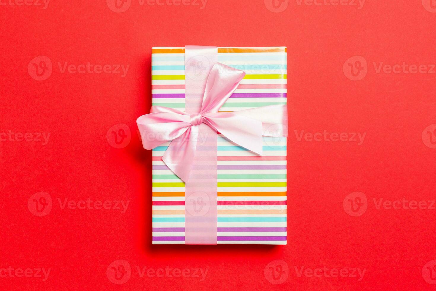 wrapped Christmas or other holiday handmade present in paper with pink ribbon on red background. Present box, decoration of gift on colored table, top view with copy space photo