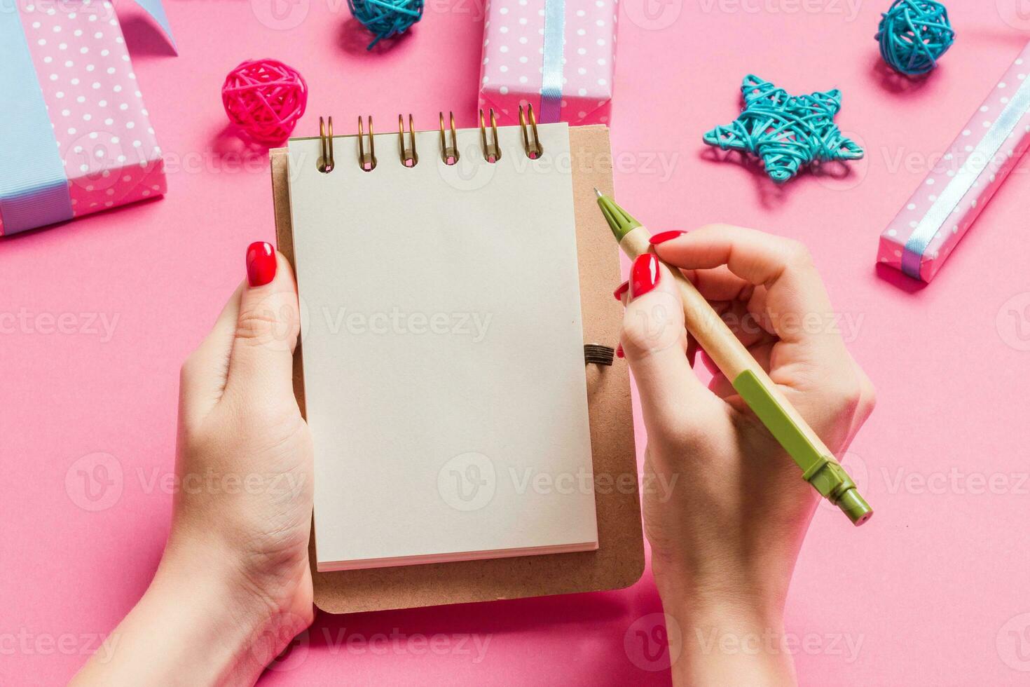 Top view of female hand making some notes in noteebok on pink background. New Year decorations and toys. Christmas time concept photo