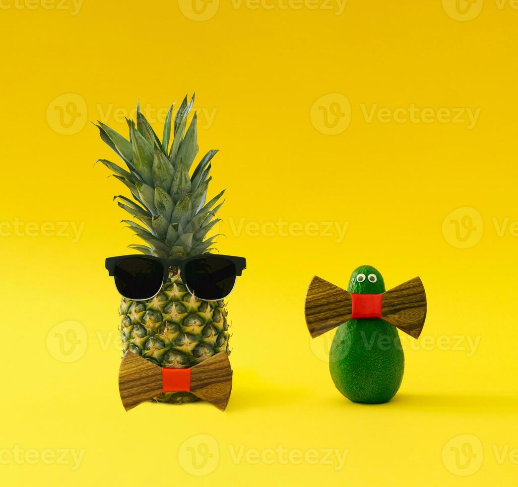 Creative and funny composition made of fresh pineapple and avocado with sunglasses and wooden bow tie on yellow background. Minimal exotic fruit concept. Trendy food idea. Tropical fruit aesthetic. photo