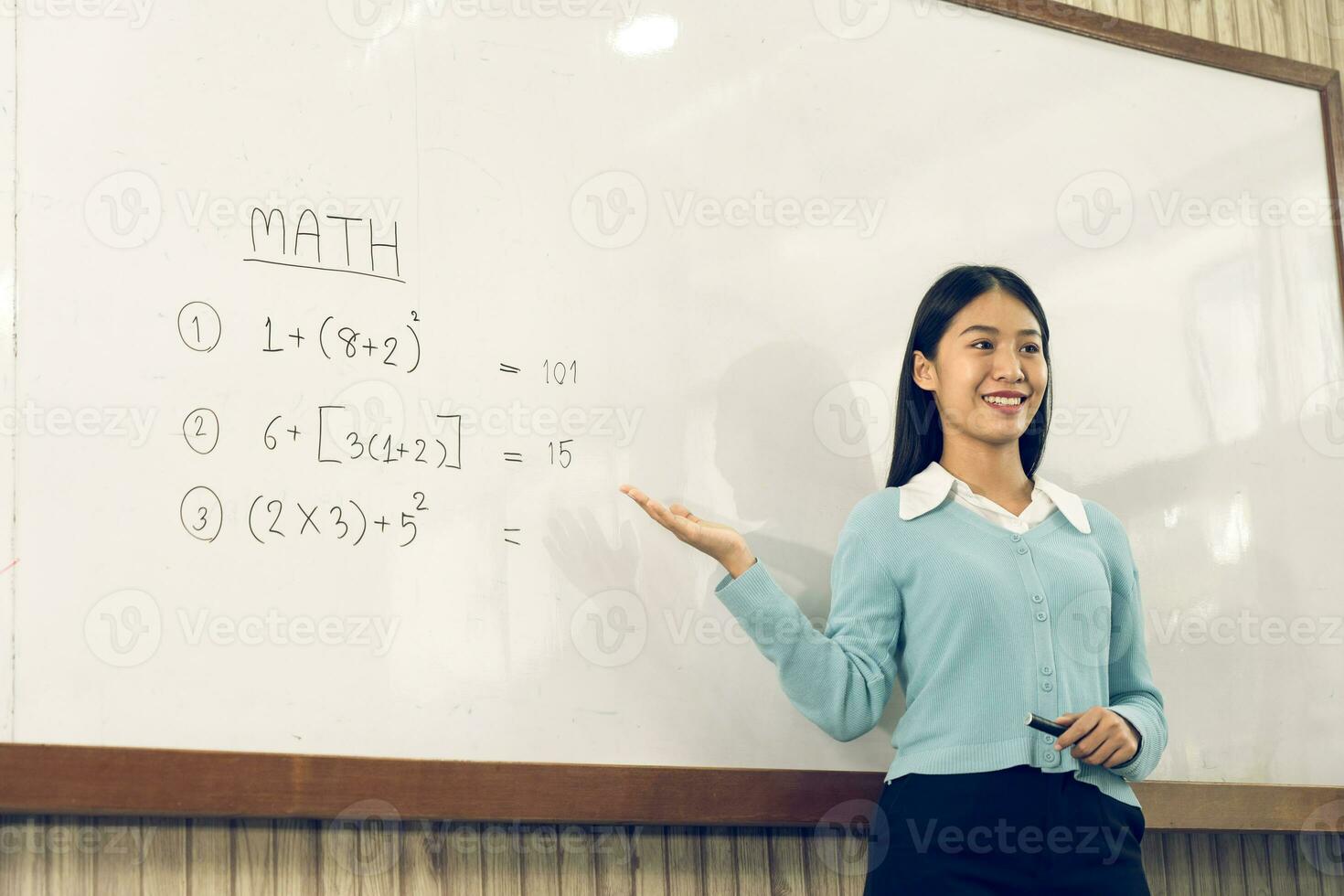Asian female teacher is teaching students at the classroom while pointing at numbers on the white board. photo
