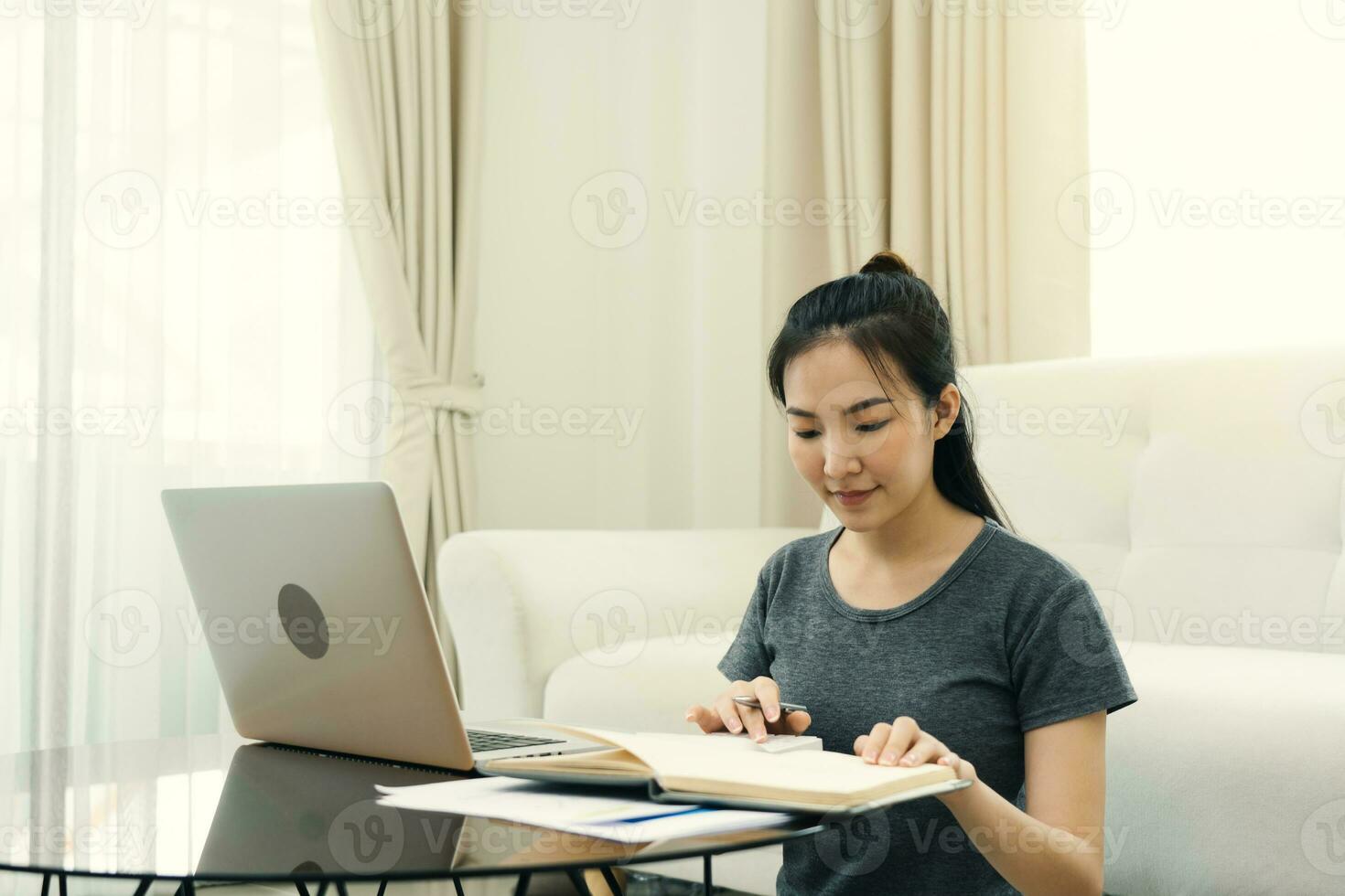 una joven asiática se sienta en la sala de estar usando una calculadora para calcular los ingresos y gastos familiares y lo escribe en su cuaderno o computadora portátil en casa. foto