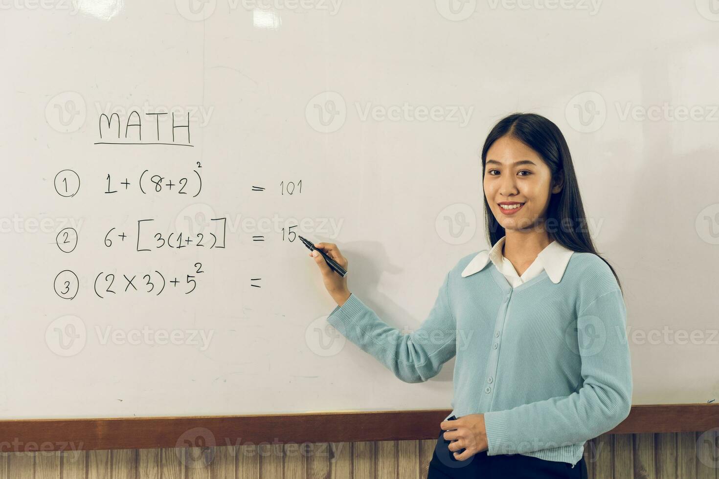 Asian female teacher is teaching students at the classroom while pointing at numbers on the white board. photo