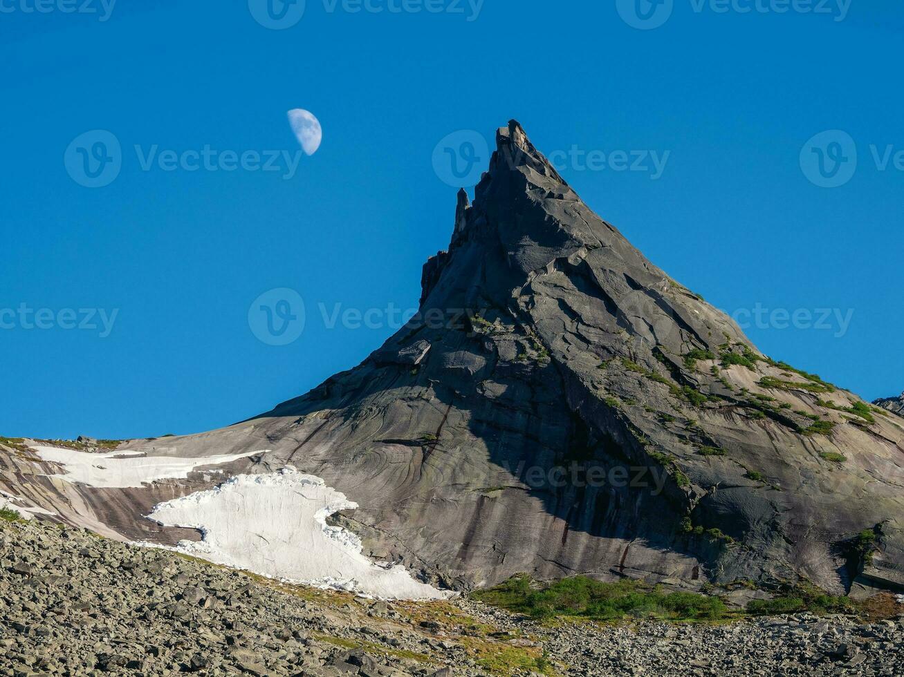 Pointed cliff with a small snow glacier, moon in the blue evening sky. Awesome scenic mountain landscape with big pointed stones in evening. Sharp rocks background. Western Sayans. photo