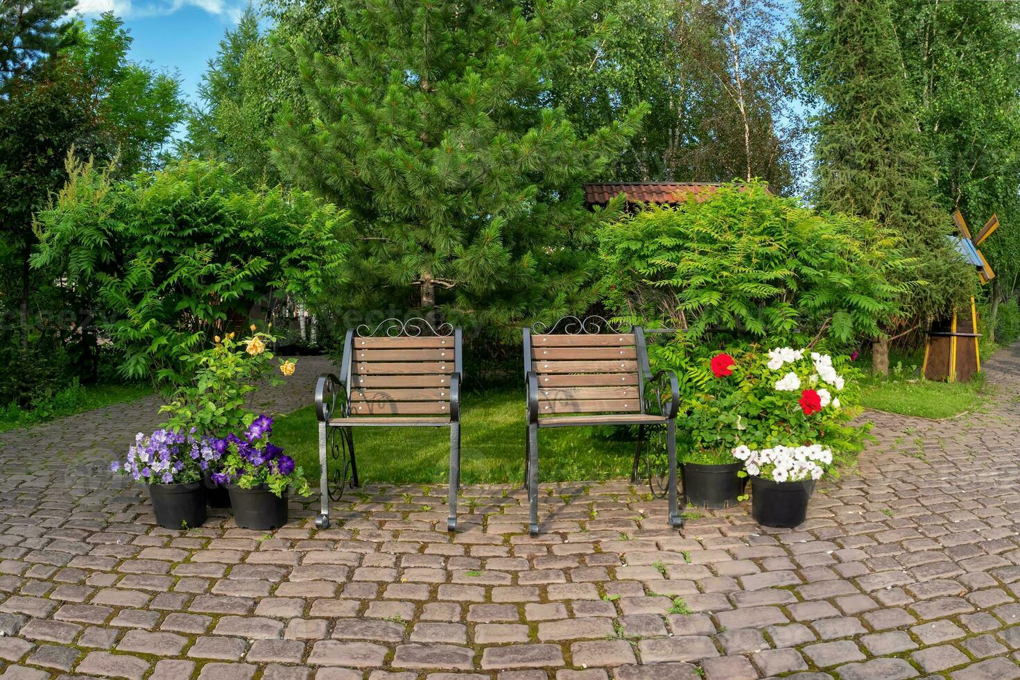 dos hierro forjado sillones en un verano jardín con albañilería de hermosa floración plantas en un Roca pavimentado pedestal paisaje diseño concepto. foto